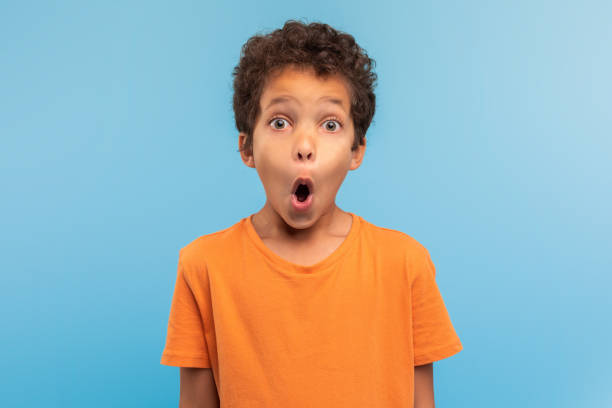 istock Surprised boy in orange t-shirt with wide eyes, blue backdrop 1994799143