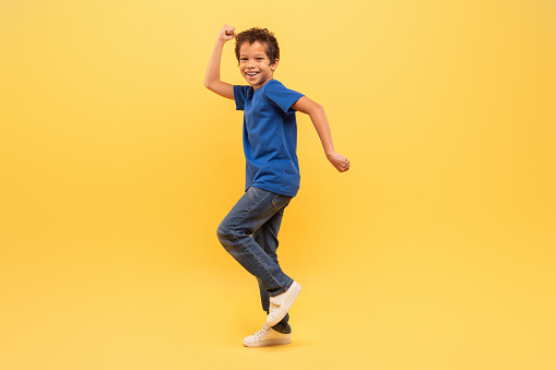 Exuberant young boy with curly hair joyfully dancing in blue t-shirt and jeans against bright yellow backdrop, expressing happiness and energy