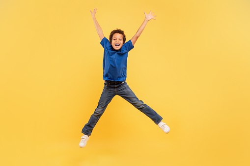 Ecstatic young boy with curly hair leaping joyfully with arms spread wide, sporting blue t-shirt and denim jeans, set against vivid yellow backdrop, embodying freedom and joy