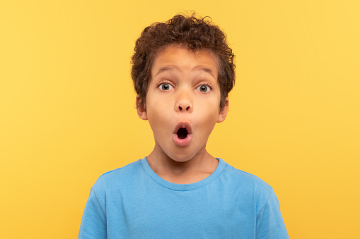 Astonished young boy with curly hair, mouth agape in surprise, wearing light blue shirt against plain yellow background, capturing moment of amazement