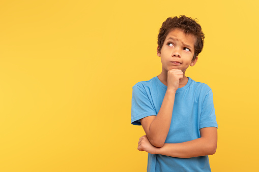 Thoughtful young boy with curly hair looking aside at copy space, touching chin in contemplation, wearing blue shirt on yellow background, embodying curiosity and deep thought