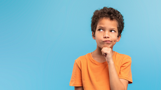 Pensive young boy with hand on chin, looking up and away at free space in deep thought against soothing blue backdrop, indicating curiosity and contemplation