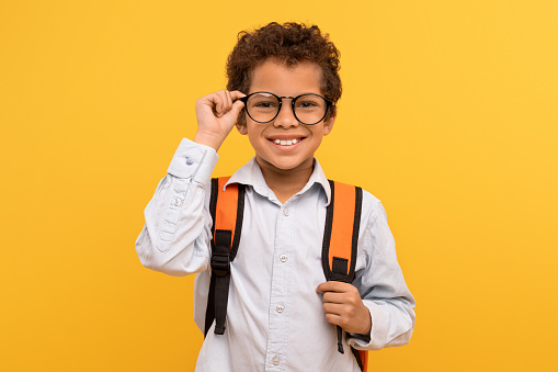 Confident schoolboy adjusting glasses, dressed in crisp white shirt with backpack, standing against yellow background, ready for day of learning
