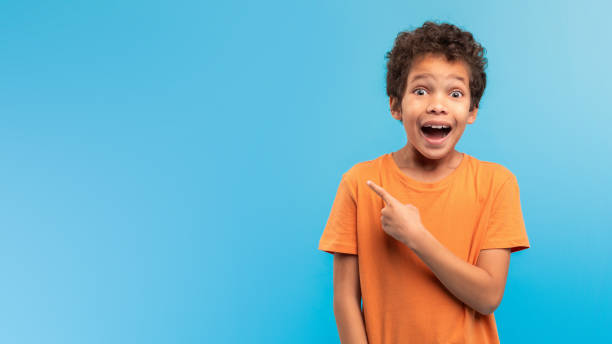 istock Astonished boy pointing at copy space, excited on blue backdrop 1994799706