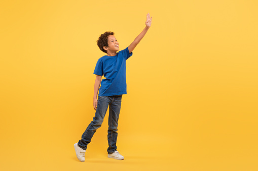 Black student boy with cheerful expression, stretching an arm to touch something unseen, clad in blue t-shirt, against bright yellow background, full of imagination