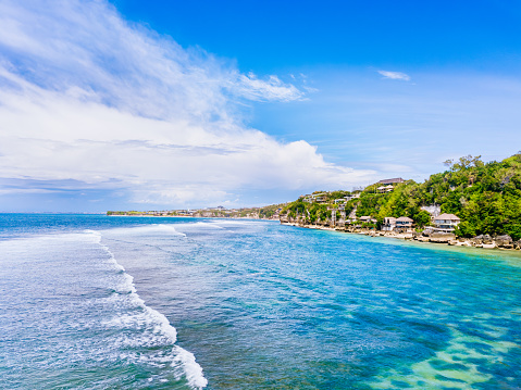 A coastal landscape dominates the view, featuring clear blue waters with gentle waves rolling towards the shore. Houses are nestled amidst lush greenery along the coastline under a sky adorned with scattered clouds. Shot taken on Bingin beach, Bukit.