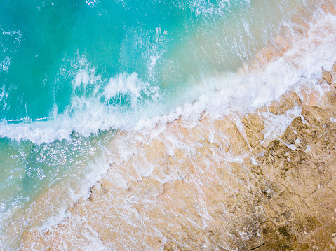 Aerial view of dynamic water and sand interaction. Waves create white foam as they meet beige sand, showcasing natural textures and patterns.