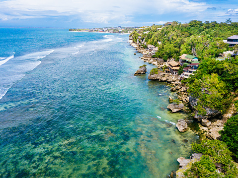 A coastal landscape dominates the view, featuring clear blue waters with gentle waves rolling towards the shore. Houses are nestled amidst lush greenery along the coastline under a sky adorned with scattered clouds. Shot taken on Bingin beach, Bukit.