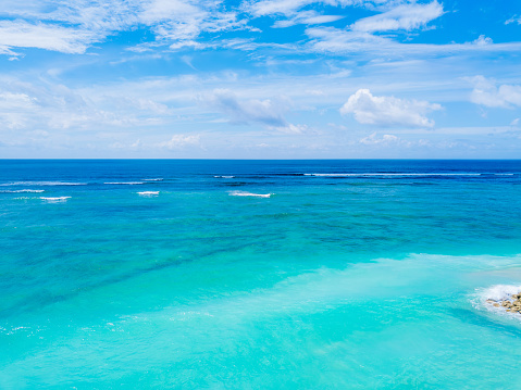 Azure ocean waters transition from turquoise to deeper shades of blue, gently rippling under the bright sky. The view captures the vastness and tranquility of the seascape, devoid of any visible land. The horizon stretches into the distance where the sky meets the sea. Shot taken in Nusa Dua, Bali.