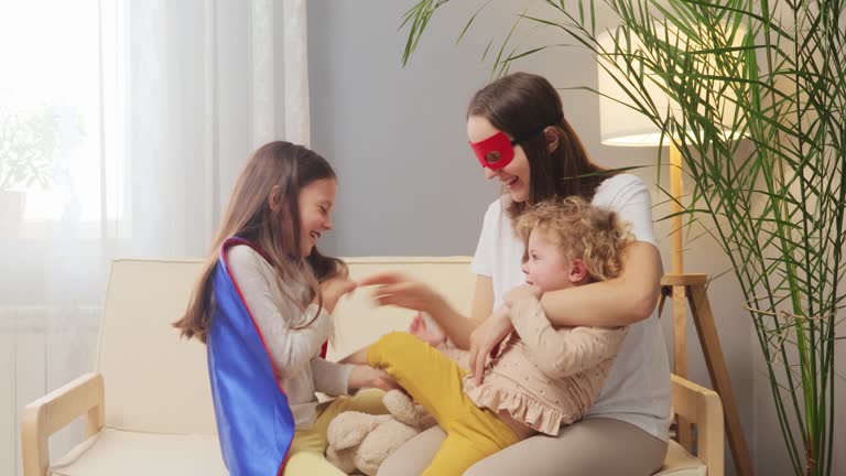 Cheerful woman wearing superhero mask playing with her daughters at home sitting on sofa in living room hugging raising fist like flying to save work laughing happily