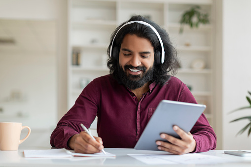 Smiling indian man enjoying music on headphones while browsing tablet and taking notes, handsome young eastern male study or working online, sitting at desk at bright home office, closeup