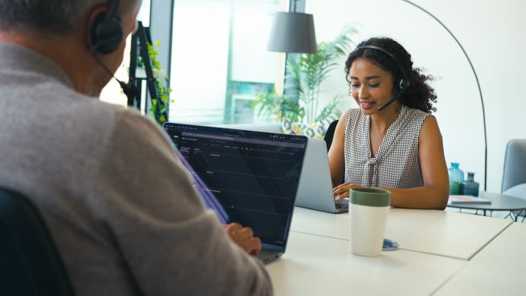 Woman in modern open plan office wearing headset sitting at desk with laptop working in call centre team taking call - shot in slow motion