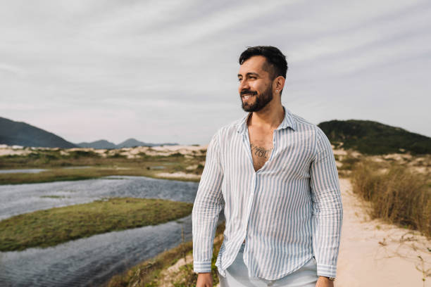 Smiling young man standing on a sandy footpath and looking at the view during a walk along a scenic coastline