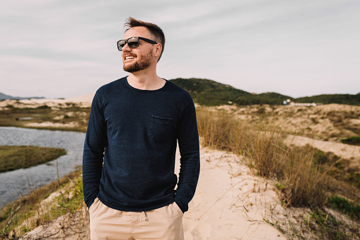 Smiling young man wearing sunglasses standing on a sandy footpath and looking at the view during a walk along a scenic coastline