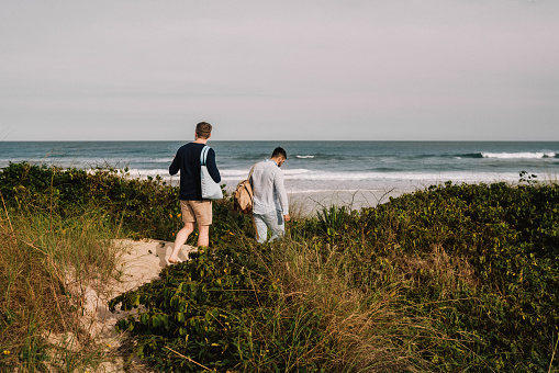 Rear view of a young gay couple walking together along a sandy path during a day by the ocean in summer