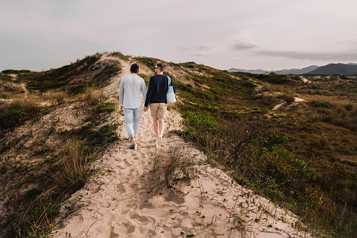 Rear view of an affectionate young gay couple walking arm and arm along a sandy path during a day by the coast