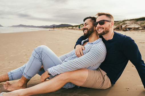 Loving young gay couple relaxing together on a sandy beach by the ocean in summer