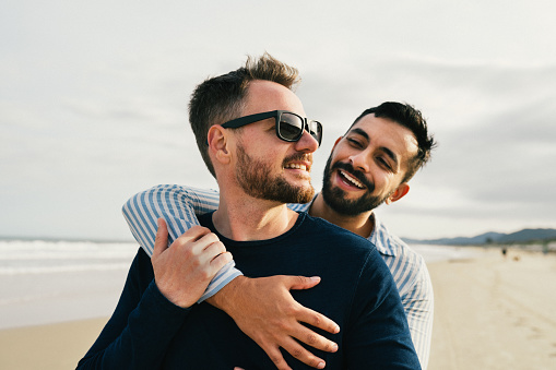 Affectionate young gay couple standing arm in arm together on a sandy beach by the ocean in summer