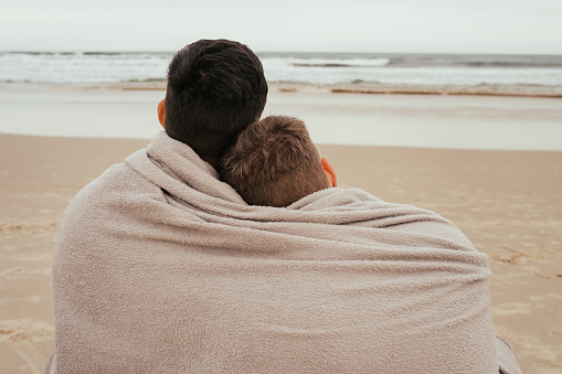 Rear view of an affectionate young gay couple looking at the view while sitting wrapped in in a blanket on a sand beach by the ocean