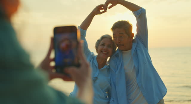 Closeup of Asian senior adult couple is photographed by a local female guide while traveling on vacation on beach enjoy sea at sunset light. Happy elderly people lifestyle after retirement.