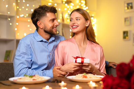 Loving man looks at his partner as she holds small gift with red ribbon, both at candlelit dinner with a warm, festive light backdrop