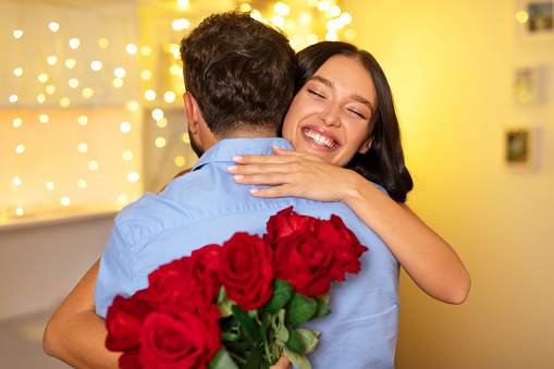 Woman with radiant smile hugs man from behind and holding beautiful bouquet of red roses, with warm bokeh lights in the background