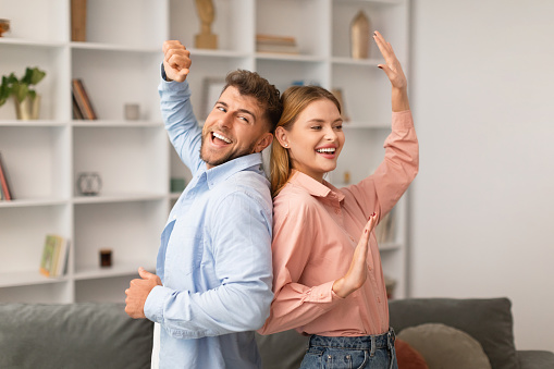 Millennial couple dancing together in living room, celebrating Valentine's Day at home. Young husband and wife smiling in love, sharing romantic and fun moment during anniversary celebration