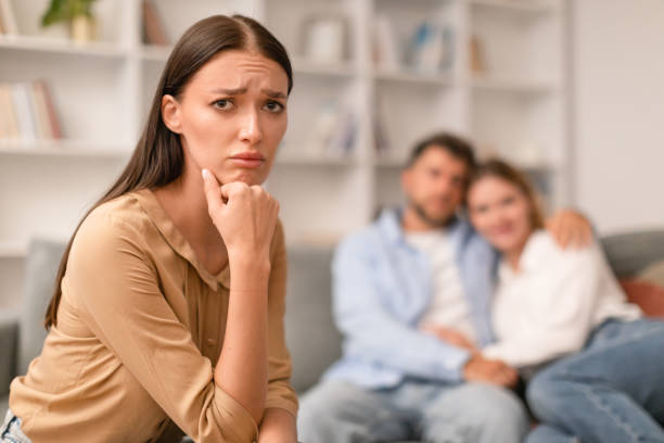Sad Woman Sitting While Her Ex Boyfriend Hugging Girlfriend Indoors Jealousy. Sad Young Woman Looking At Camera While Her Ex Boyfriend Hugging New Girlfriend Sitting On Couch Indoors. Love Triangle And Envy In Friendship With Married Friends. Selective Focus jealous ex girlfriend stock pictures, royalty-free photos & images
