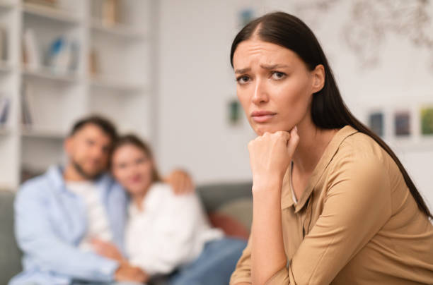 woman suffering from jealousy while man hugging another lady indoor Complicated love triangle. Unhappy young woman suffering from jealousy while man hugging another lady, looking sadly at camera, feeling tension of infidelity in modern living room. Shallow depth jealous ex girlfriend stock pictures, royalty-free photos & images