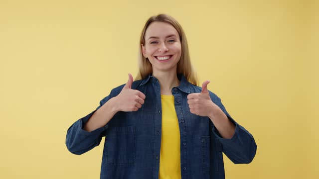 Portrait of cheerful female wearing denim jacket making thumb up sign and smiling over isolated yellow background. Pretty woman looking at camera and showing support and respect gesture indoors.