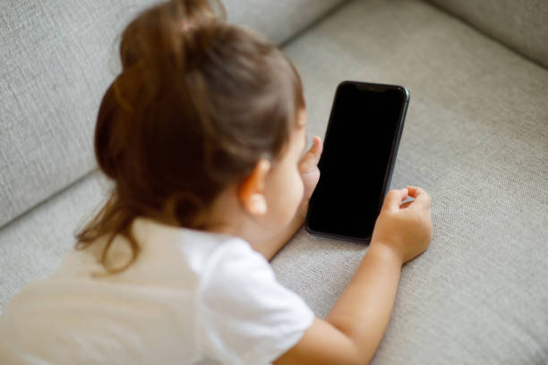 istock Little girl relaxing on comfortable couch and using smartphone with blank screen 1994947692
