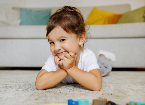 Little Cutie. Adorable Cheerful Toddler Girl Lying On Floor And Smiling At Camera, Happy Preschool Female Child Resting Head On Hands, Relaxing On Carpet In Cozy Living Room Interior, Closeup