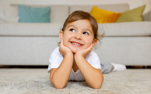 Cheerful cute little girl daydreaming on the floor at home, adorable toddler female child lying on carpet in living room, resting head on hands, smiling and looking away, copy space