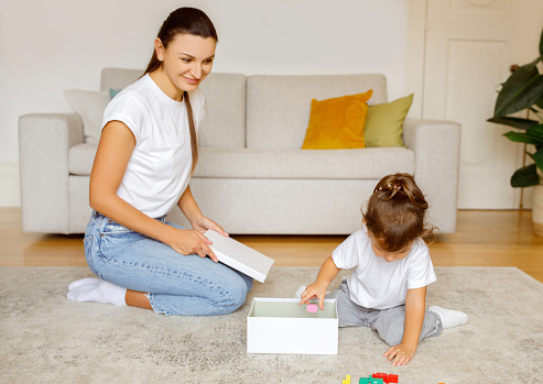 Happy mother watching her daughter discovering contents of surprise gift box during playful and educational moment on their living room floor, mom and female child playing together at home