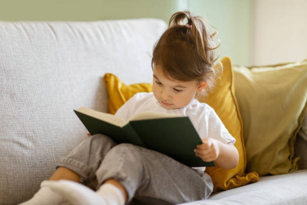 istock Cute little girl reading book while sitting on couch at home 1994948273