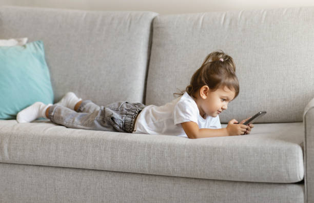istock Cute toddler girl using smartphone while lying on comfortable sofa at home 1994948278
