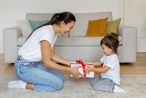 Excited mother presenting white gift box with red ribbon to her joyful preschool daughter, creating a moment of happiness in a comfortable living room, happy girl taking present from mom and smiling