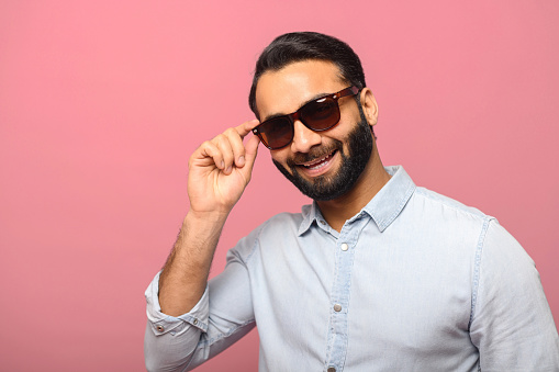 Headshot of handsome positive bearded man wearing sunglasses and blue casual shirt posing over pink background, looks at the camera, flirting