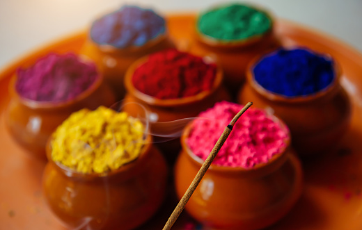 Burning and fuming aroma stick against circle of Holi powder in clay cups. Happy Holi greeting card and background.