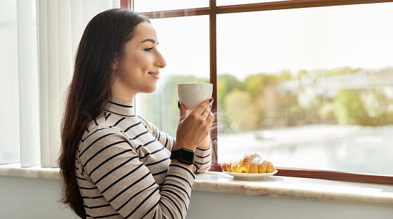 Contented cheerful middle eastern young woman enjoying a peaceful moment, sipping coffee and gazing out a sunny window, with a fresh croissant waiting on the marble windowsill