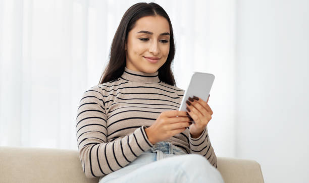 Glad young arab woman typing by smartphone at sofa, watch video, read message