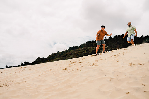 Laughing teenage boy riding a sandboard down a dune with his father running behind him during a beach vacation in summer