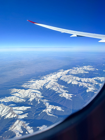 Stock photo showing the wing of a plane pictured through the airplane window with views of a dramatic snowy mountain range and blue sky pictured through an airplane window.