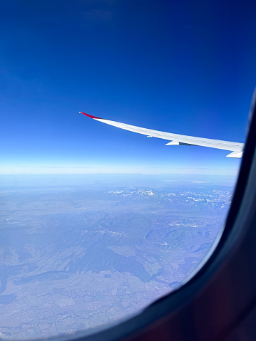 Stock photo showing the wing of a plane pictured through the airplane window with views of a dramatic snowy mountain range and blue sky pictured through an airplane window.