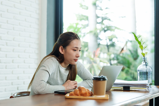 Beautiful young Asian woman sitting at table in a cafe After stress of verifying the store's account Drink coffee in afternoon to keep yourself from feeling sleepy. Croissants were placed on table.