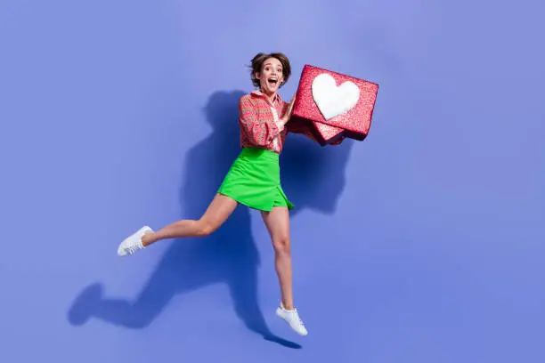 Photo of Full length photo of charming ecstatic girl wear green skirt jumping with social media like in hands isolated on blue color background