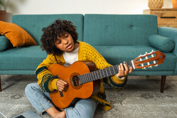 istock African American woman learning guitar watching tutorial on laptop at home. Black girl playing guitar singing song learning online music lessons, Artistic woman playing acoustic guitar learning chords 1995099114