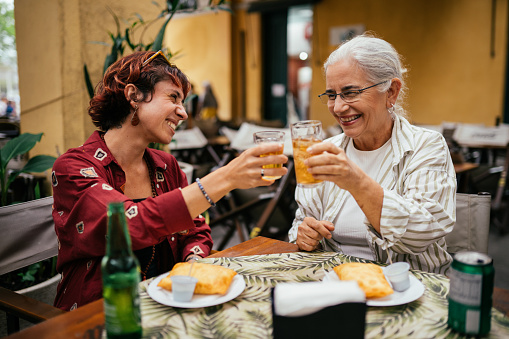 Smiling mature woman and her adult daughter toasting with drinks while having lunch outside at a table in a restaurant courtyard in Municipal Market in Florianopolis, Brazil