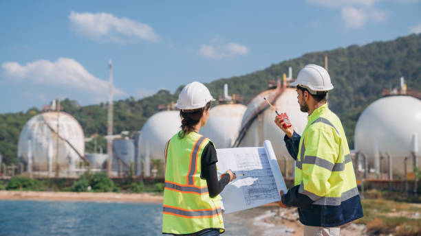 istock Two engineers in safety gear with blueprints discussing work in front of industrial storage tanks on a clear day. 1995111909