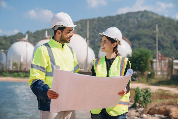 istock Two engineers in safety gear with blueprints discussing work in front of industrial storage tanks on a clear day. 1995112962